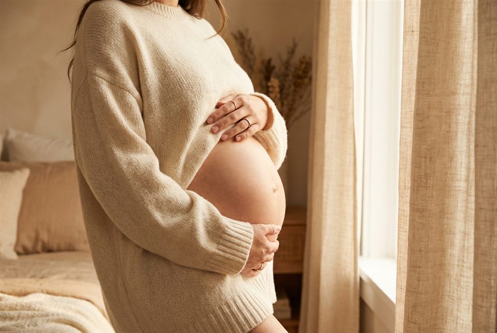 Expecting mother in a cream sweater gently cradling her belly in soft window light